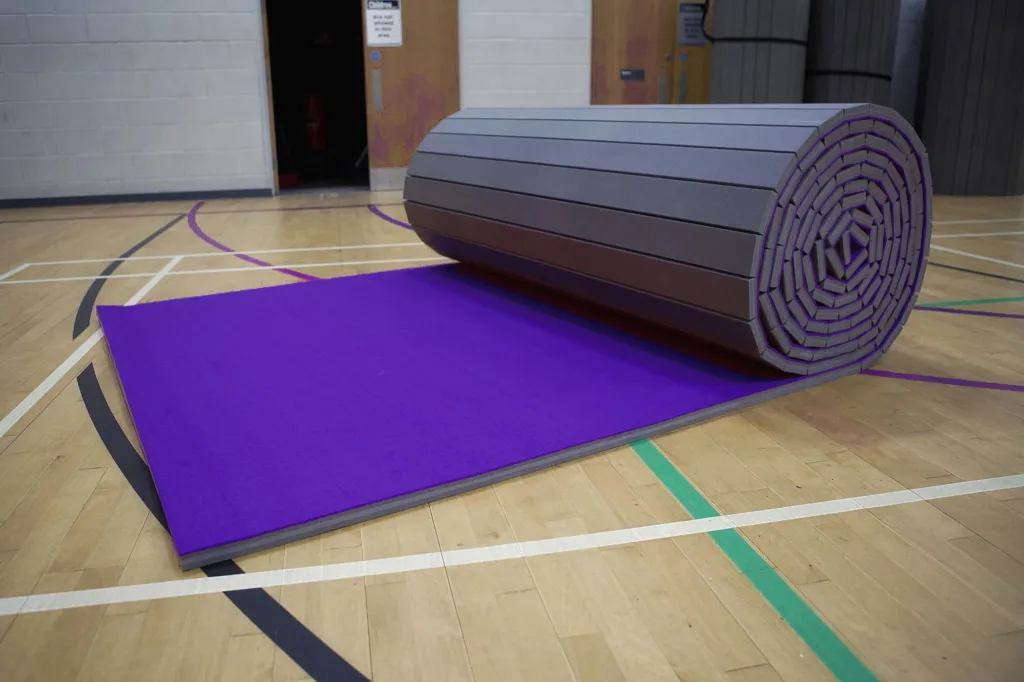 Purple carpet roll-out mat partially unrolled on a gymnasium wooden floor, revealing its segmented grey XPE foam core.