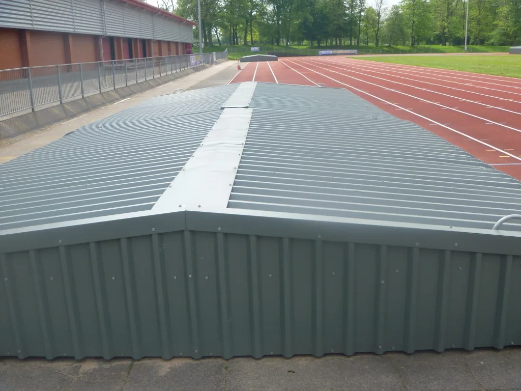 Steel wheelaway sandpit cover with corrugated epoxy-painted cladding in two sections beside a red running track.