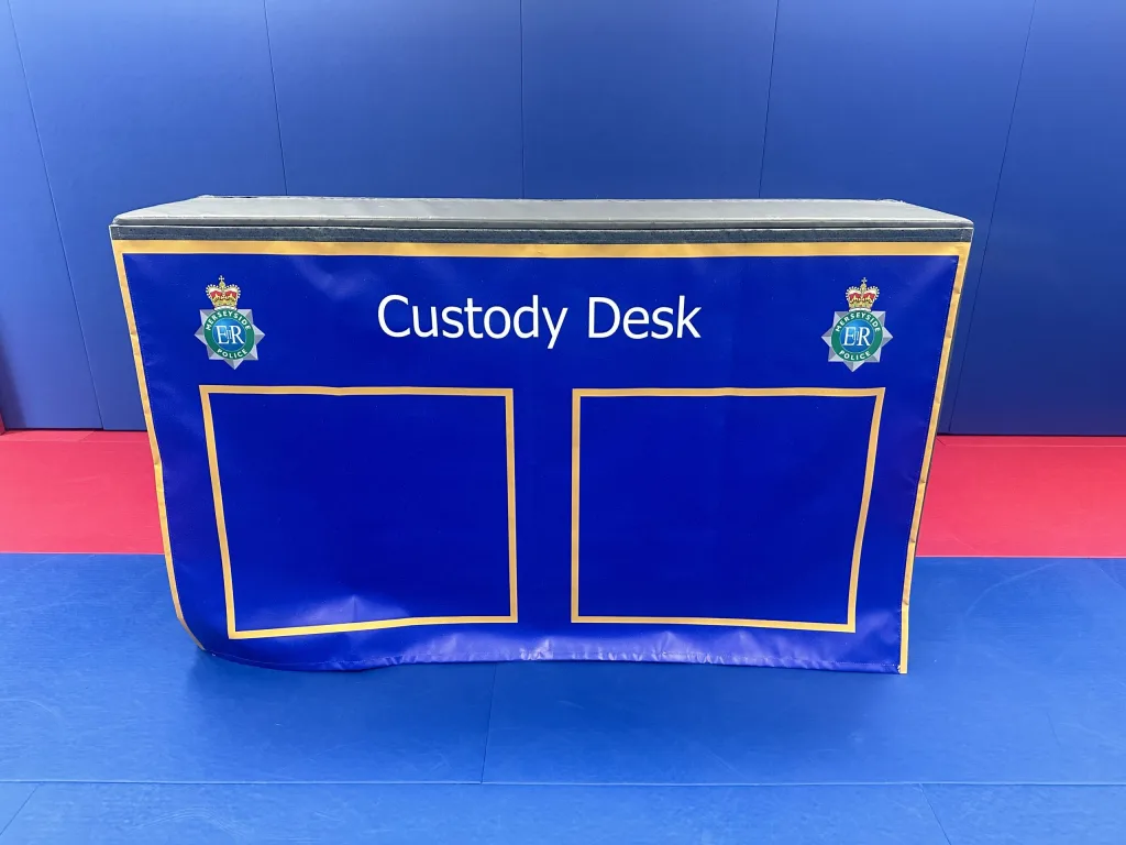 Blue multi module training block labeled Custody Desk with two gold-outlined panels and police crests, set on blue and red mats.