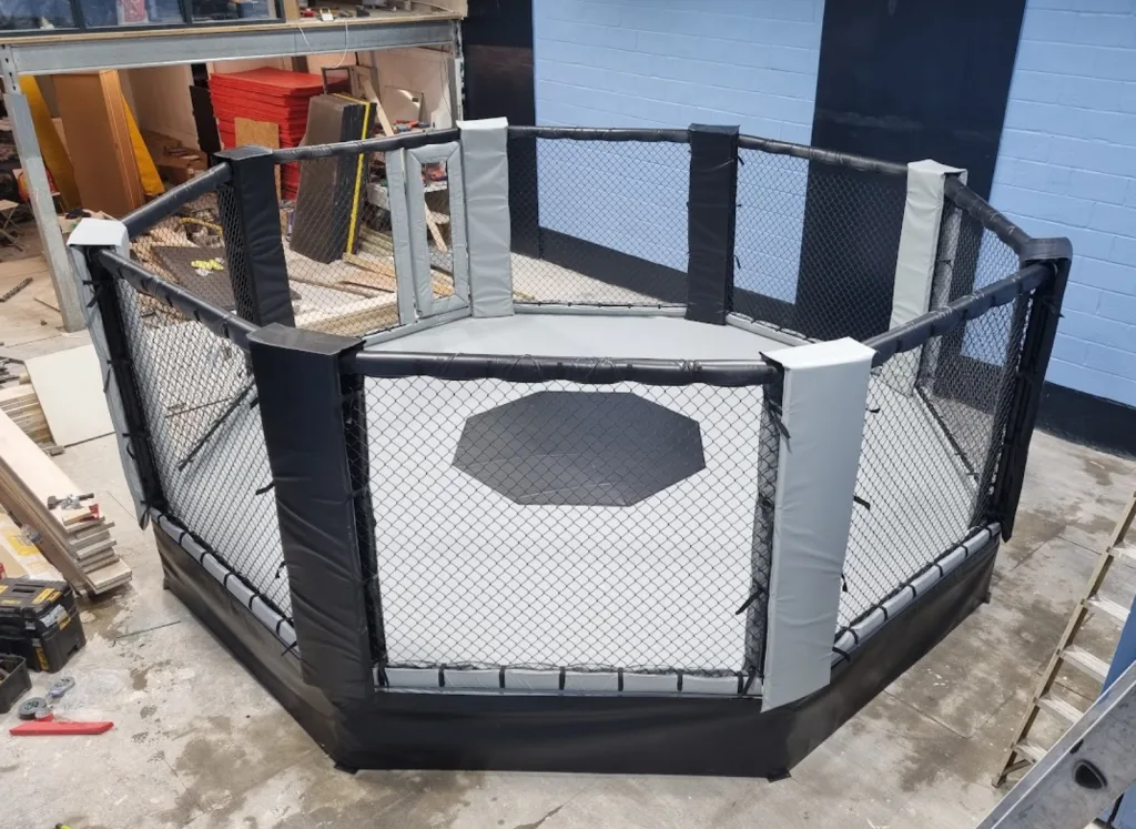 Octagonal platform training cage with a raised steel deck, powder-coated frame, chain-link sides, and padded corner posts.