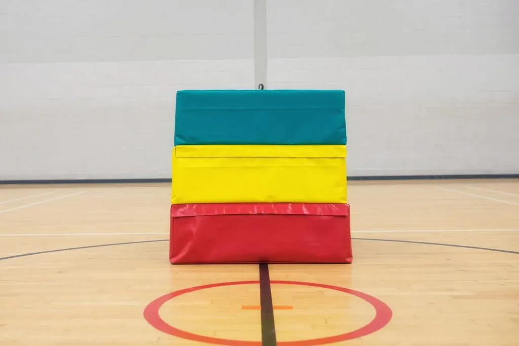 Three-layer vaulting box in a gym, top teal, middle yellow, bottom red, with PVC-coated nylon cover and carry handles.
