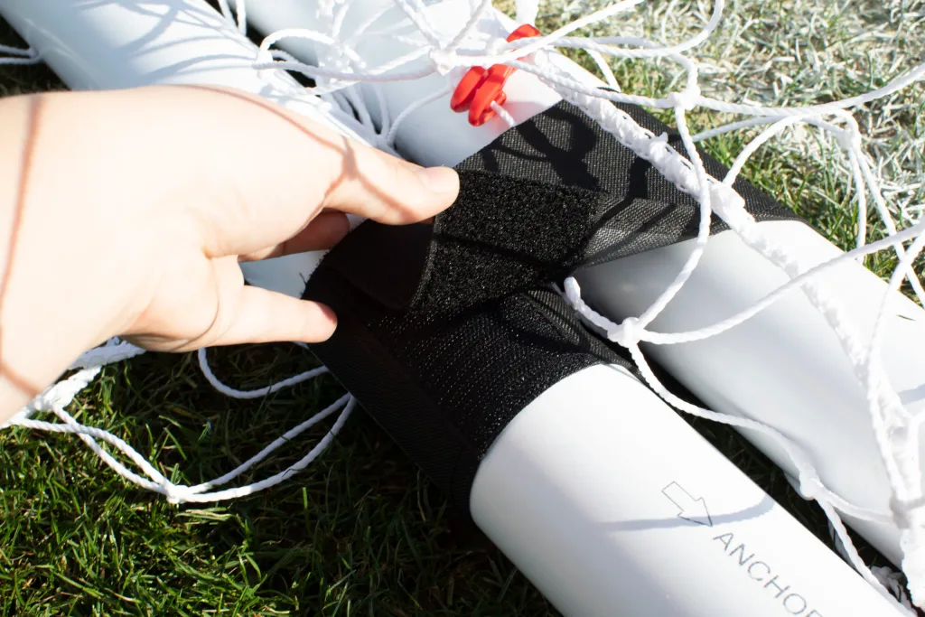 Hand fastening black Velcro strap around white goal frame tube labeled ANCHOR with white netting on grass.