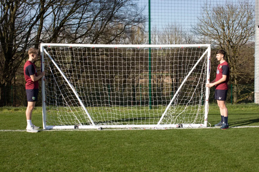 Two men stand at either end of a white fold-away soccer goal with square mesh netting on a grass training pitch.