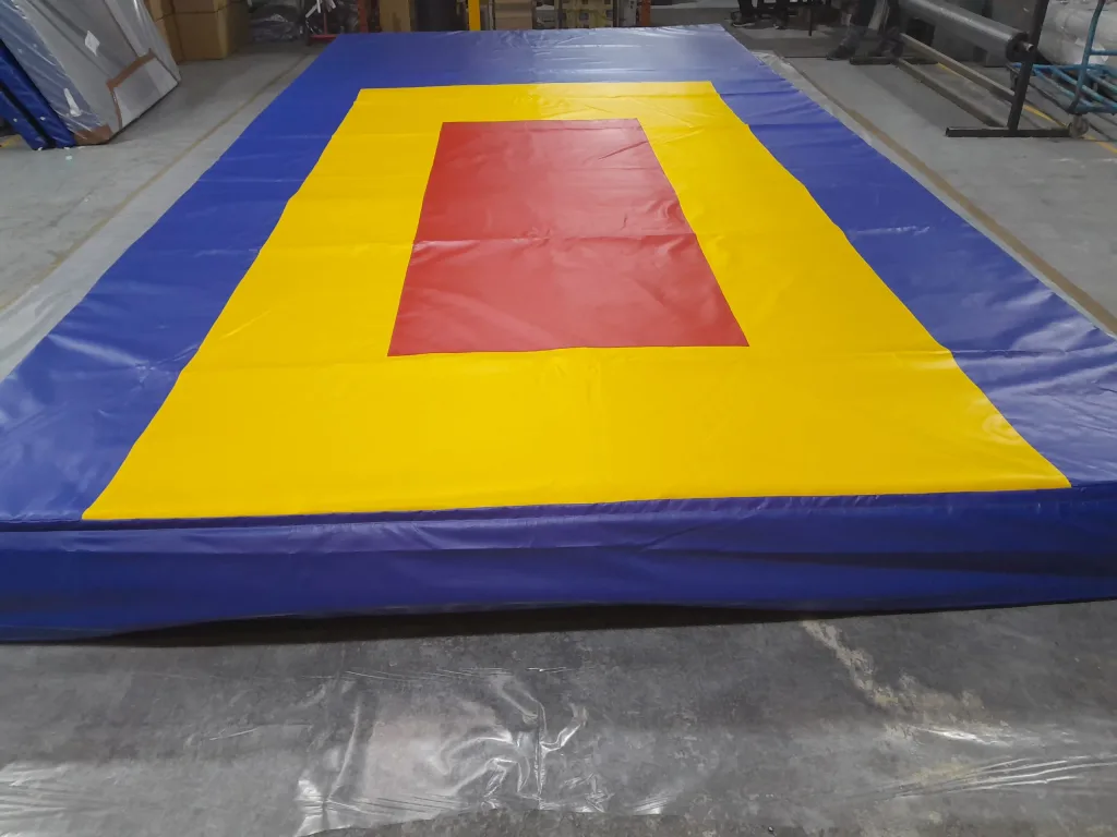 Large rectangular gymnastics landing mat with blue outer border, yellow landing area and central red rectangle on a concrete warehouse floor.