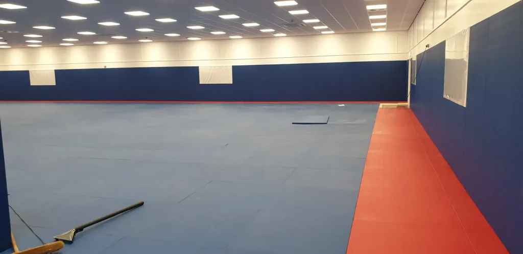 Large sports hall with blue tatami PVC judo mats, red perimeter strip, blue padded walls and overhead ceiling lights.