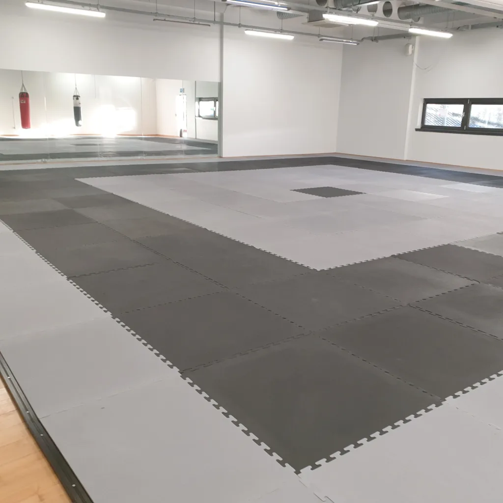 Grey and black interlocking jigsaw mats cover a training room floor, with a mirrored wall and hanging punching bags visible.