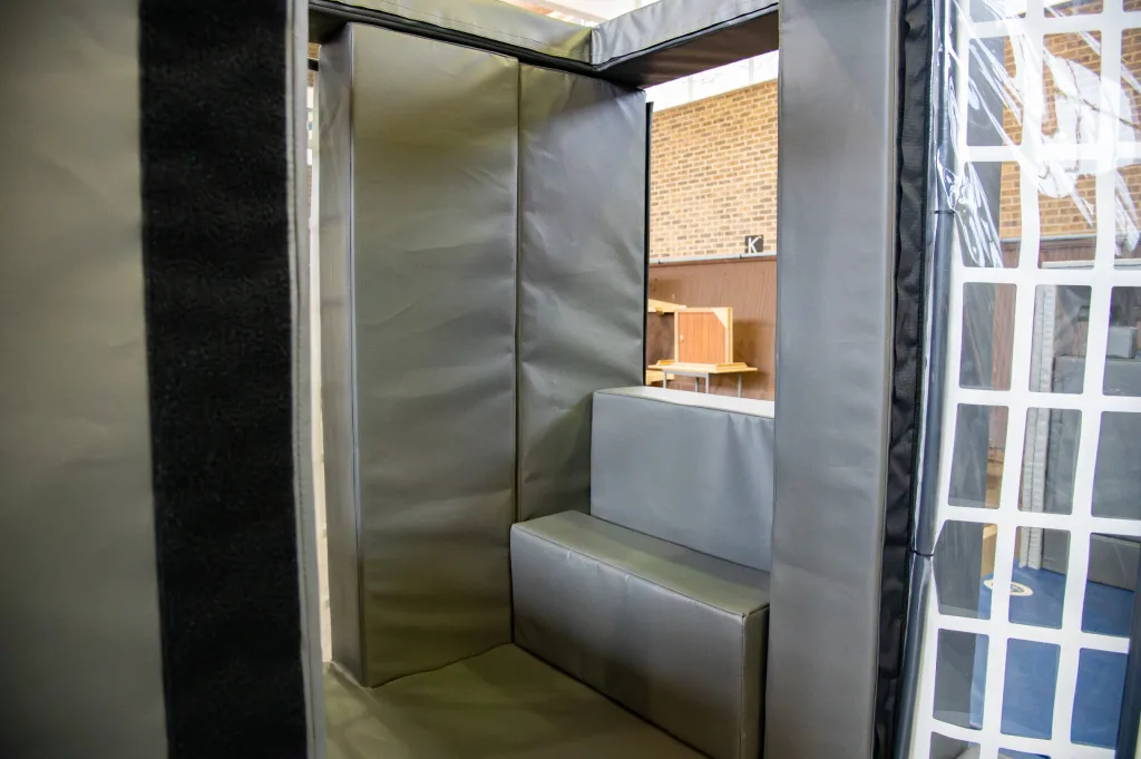 Padded confined-space training cage interior with reinforced foam and heavy-duty PVC cover, showing bench-style foam seats and wall padding.