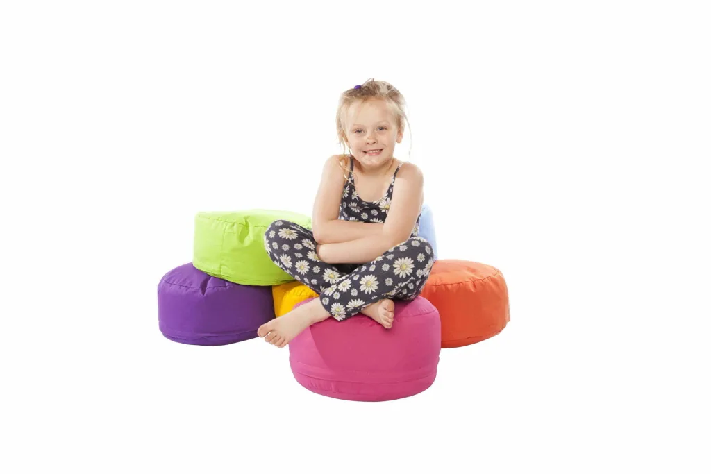 Child sitting cross-legged on a pink cotton round bean pod, smiling and surrounded by purple, lime green, yellow, orange and light blue pods.