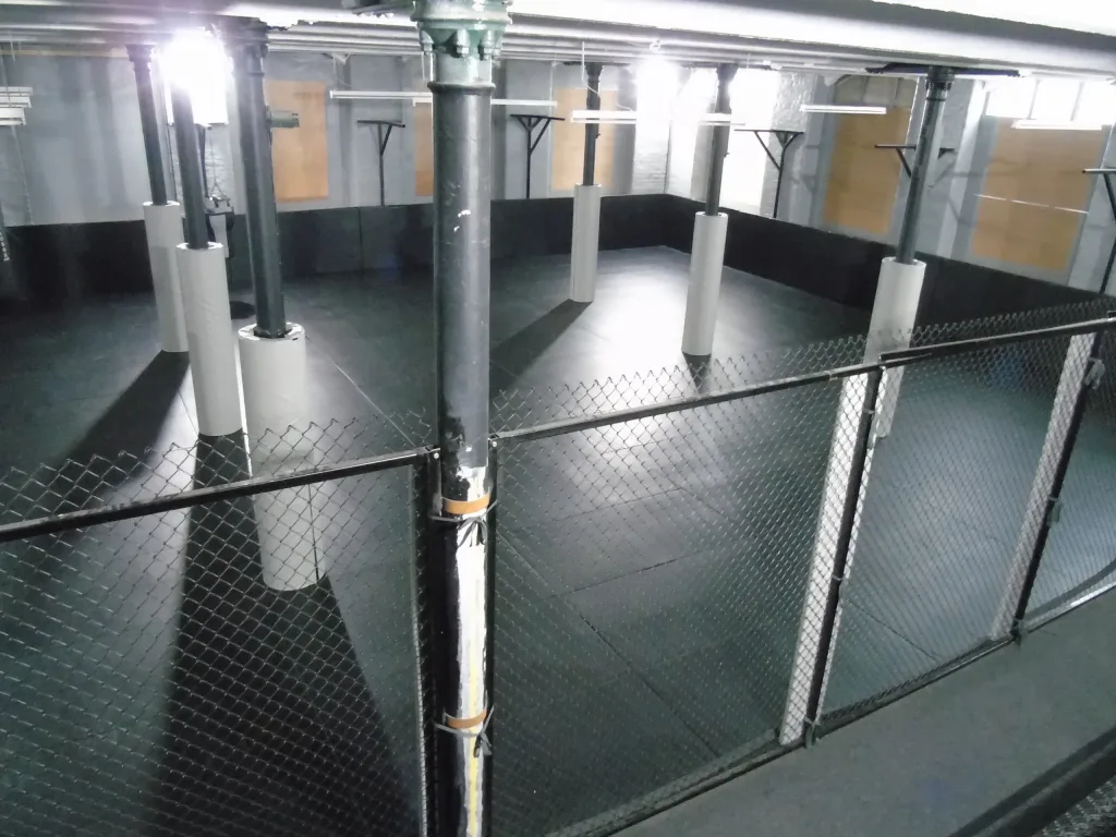 Raised view over a fenced indoor training area with black heavy-duty polyester PVC mats covering the floor and light grey padded support columns.