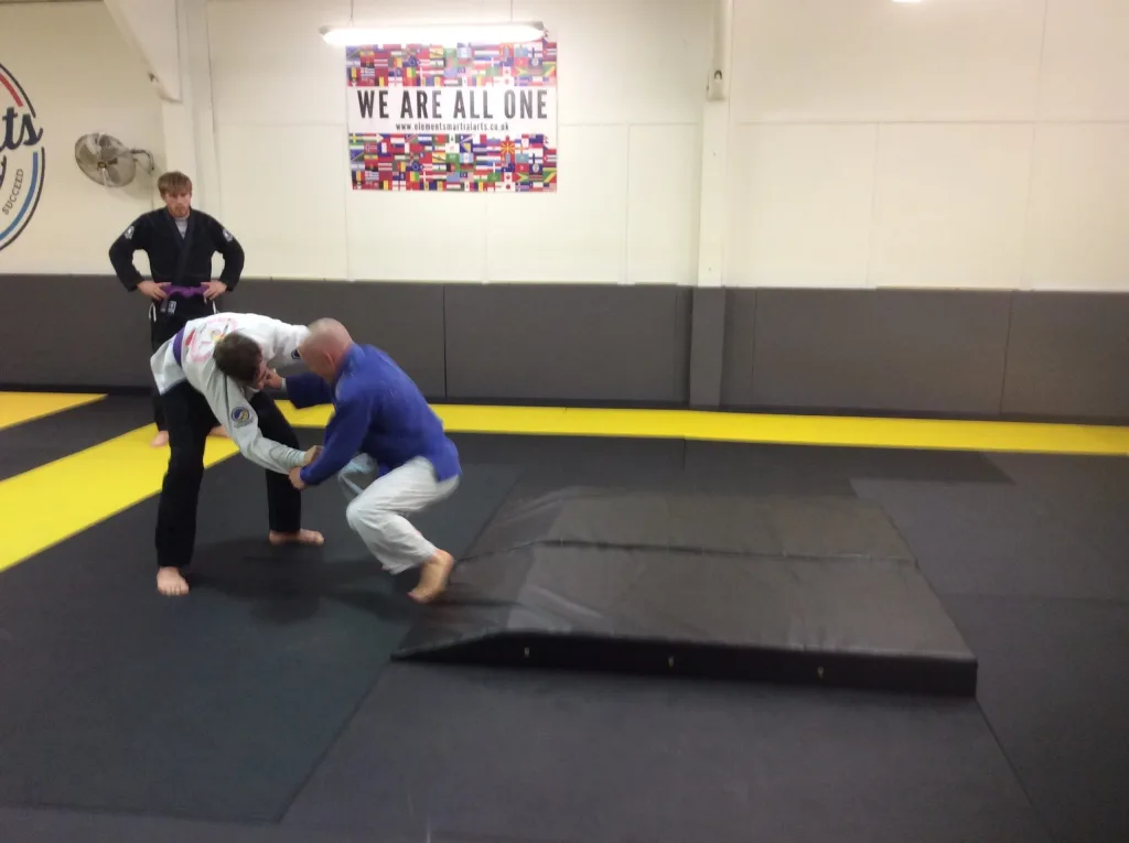 Black PVC slam mat with a chamfered end on a martial arts floor, two people practicing throws nearby and a wall sign reading WE ARE ALL ONE.