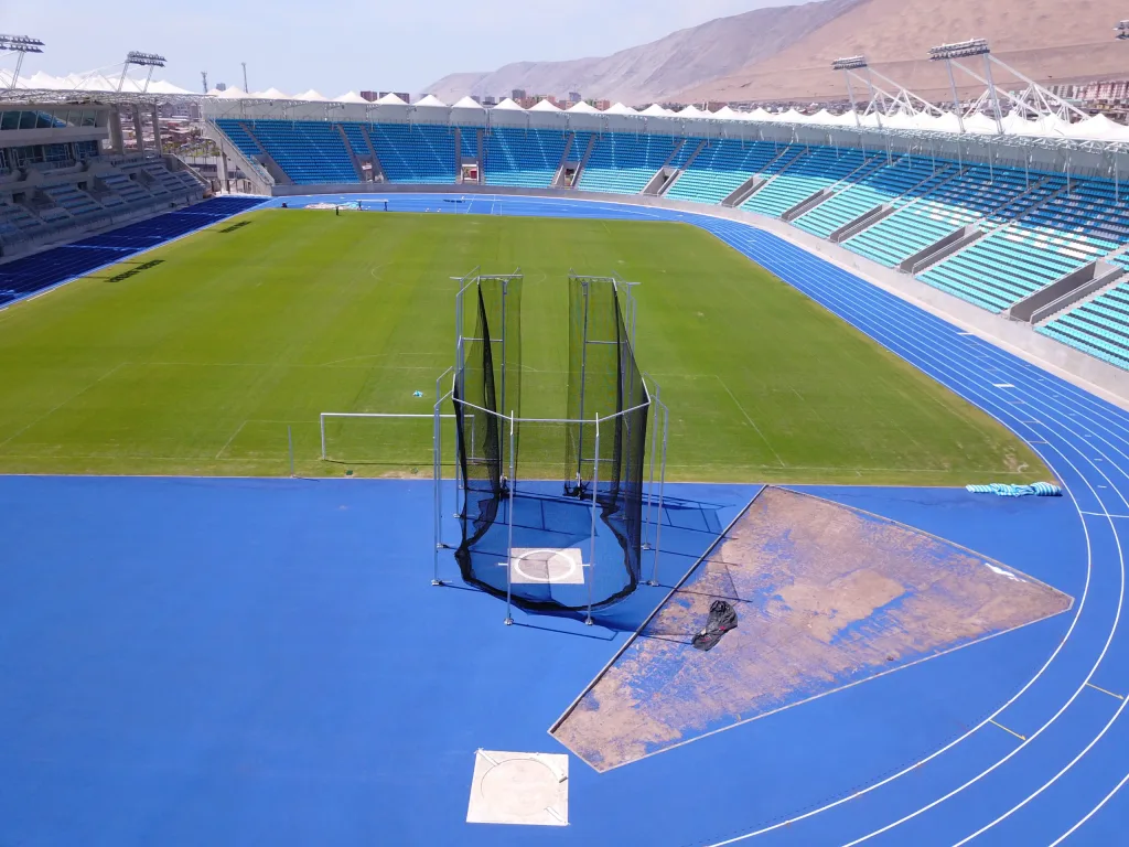 IAAF steel hammer/discus cage with black netting on galvanised steel pillars set on a blue track beside a green field in an empty stadium.