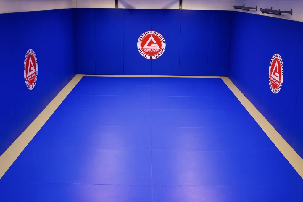 Royal blue martial arts mats with a beige border and matching wall pads, three red circular logos reading Brazilian Jiu-Jitsu Gracie Barra Belfast, N. Ireland.