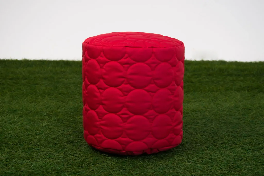 Padded red cylindrical child bean pod with quilted circle pattern placed on green artificial grass against a white backdrop.