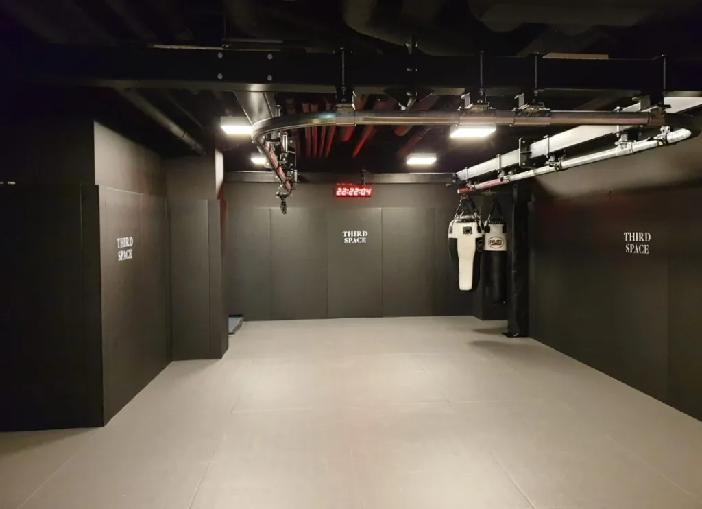 Empty MMA training room with light grey foam mats, black padded walls labeled THIRD SPACE, hanging punching bags and overhead lights.