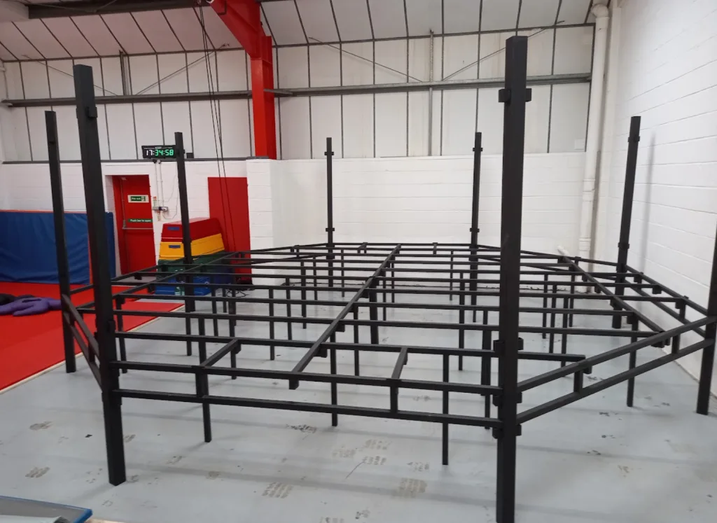 Platform training cage with steel powder-coated frame and raised deck framework in a gym, digital clock displaying 17:34:58.