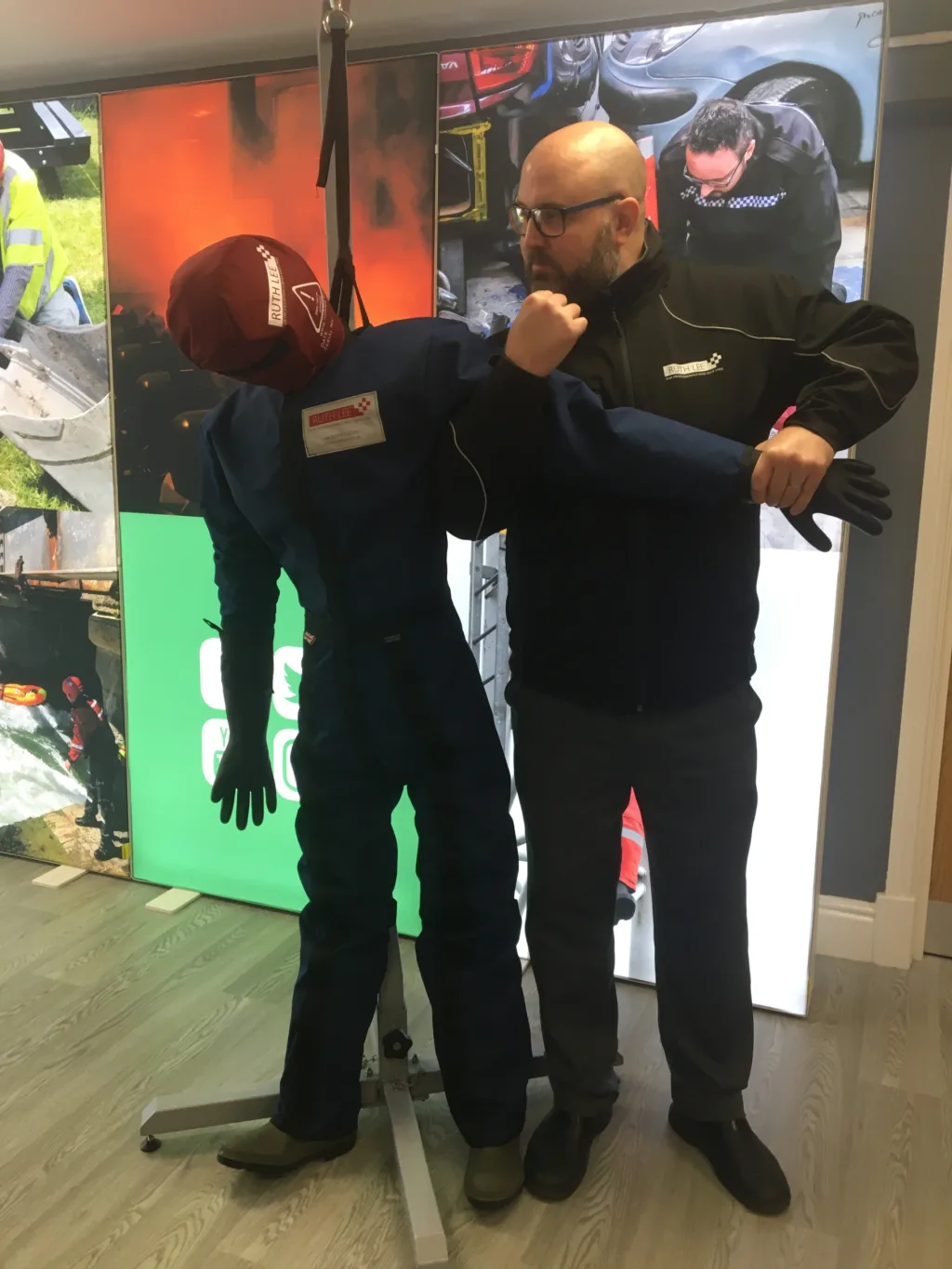 Adult PMVA manikin in blue overalls and red hood suspended on a stand while a trainer demonstrates a restraint hold.