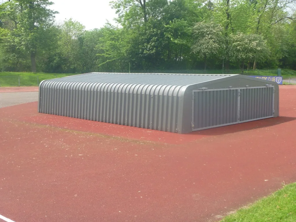 Gray corrugated galvanised steel high jump wheel-away cover lowered flush to the ground on a red track with trees behind.