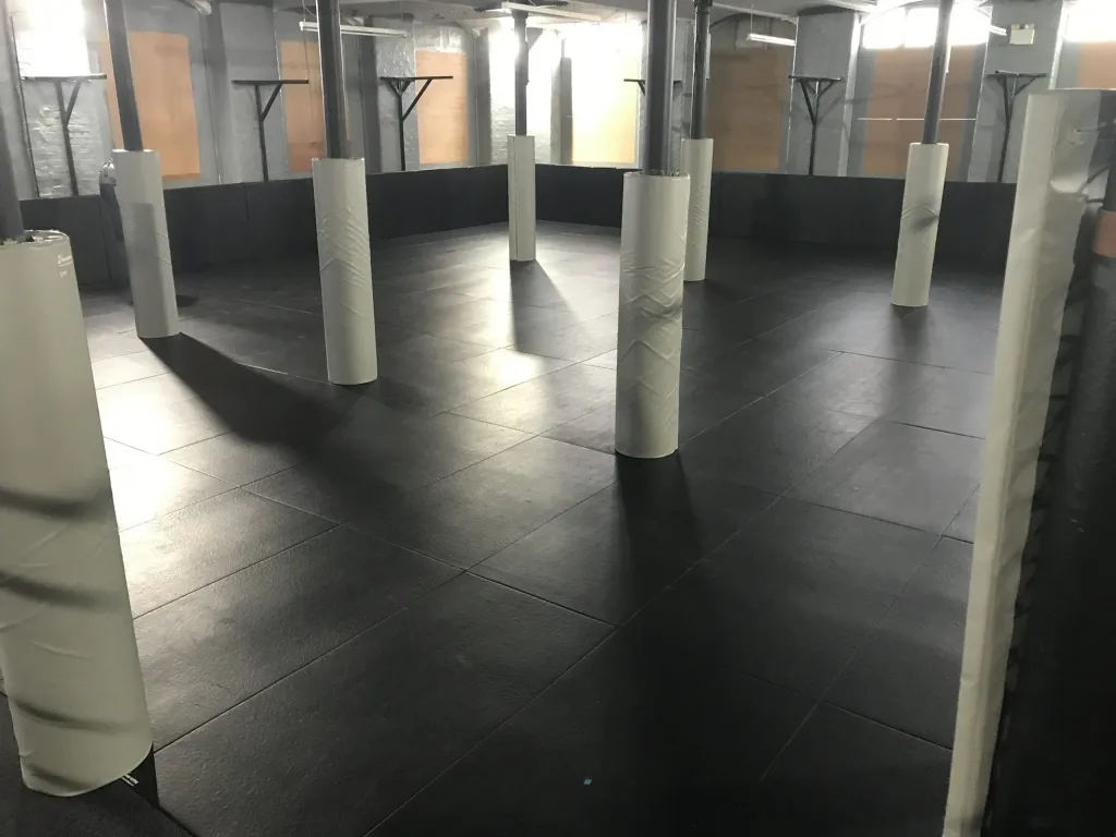 Black polyester PVC mats cover a training room floor with white padded column protectors around metal support pillars.