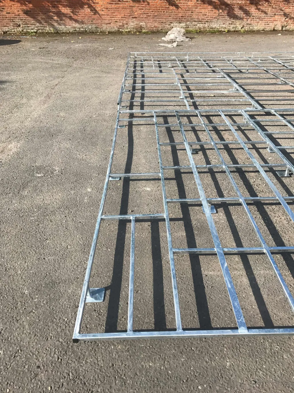 Galvanised steel raised platform grid for pole vault laid on asphalt with a brick wall in the background.
