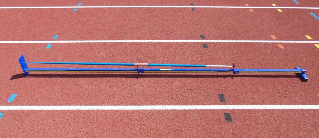Blue metal javelin gauge with a long measuring bar and sliding centre-of-gravity marker resting on a red running track.
