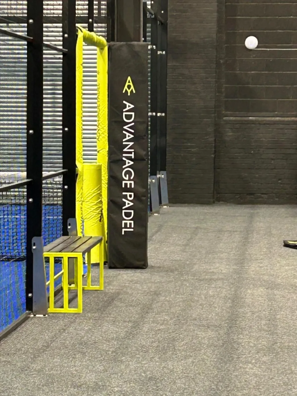 Black padded post cover labeled ADVANTAGE PADEL stands beside a yellow-wrapped post and small yellow bench on an indoor padel court.