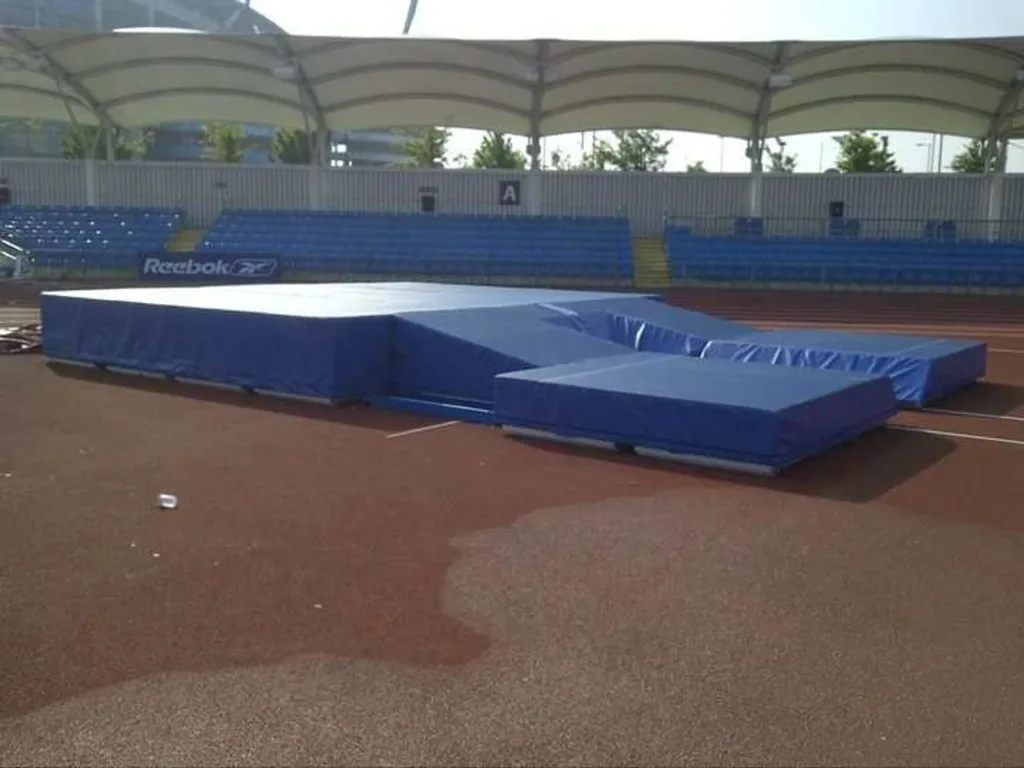 Large blue competition pole vault landing mats with reinforced PVC-coated polyester covers on a stadium track under a white canopy.