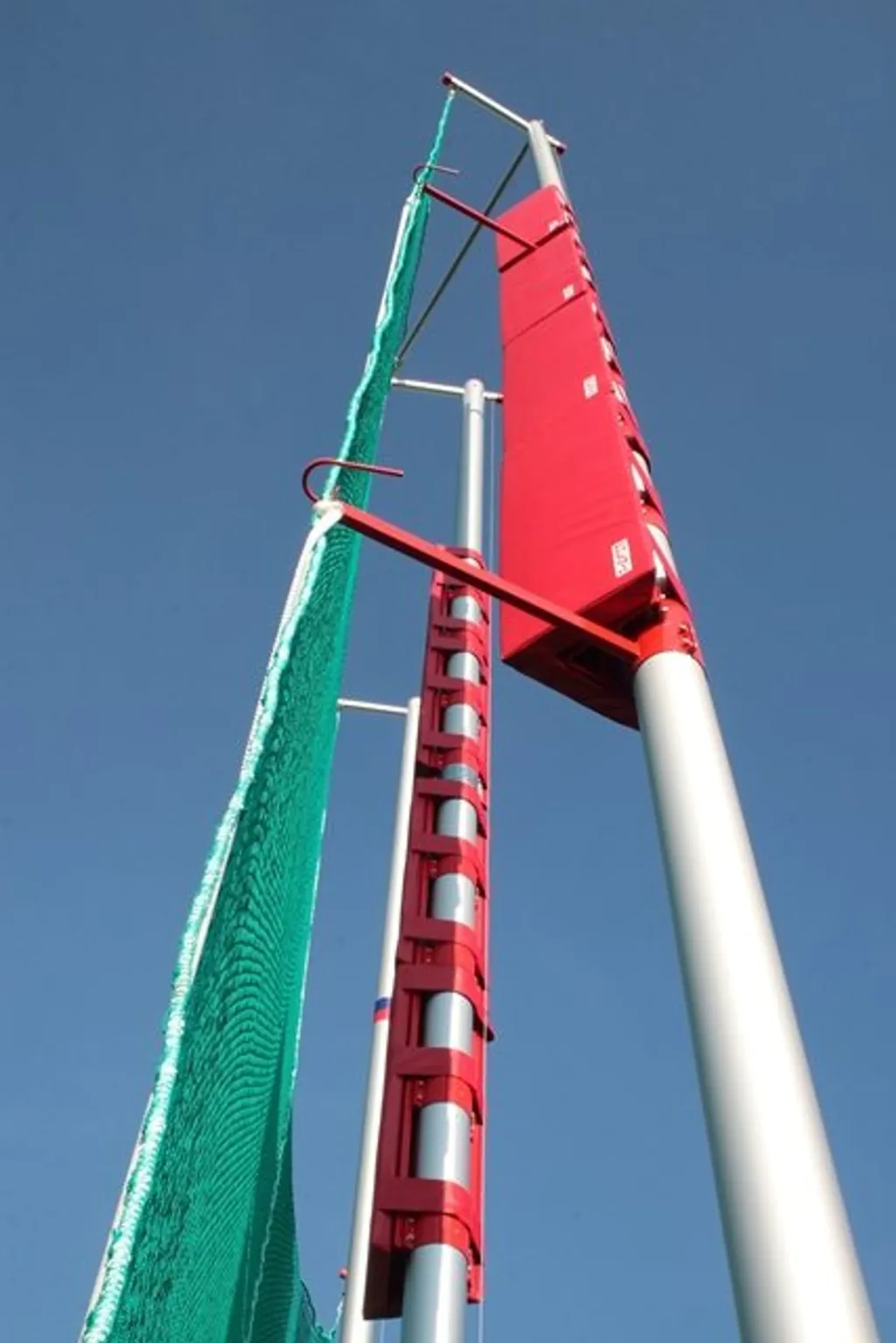 Aluminium discus and hammer cage poles with red padded supports and a green safety net reaching upward against a clear blue sky.