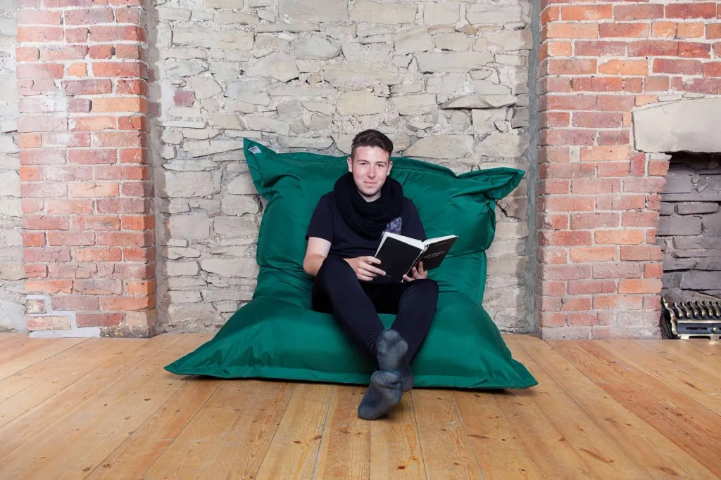 Teen sitting on a green polyester floor cushion reading a book in front of an exposed stone and brick wall.