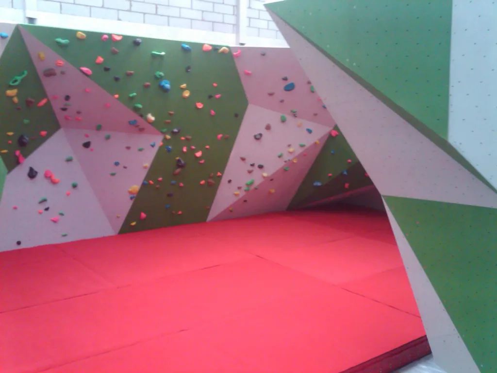 Thick red carpeted foam bouldering mats covering the floor beneath angled green and pale grey climbing walls dotted with colorful holds.