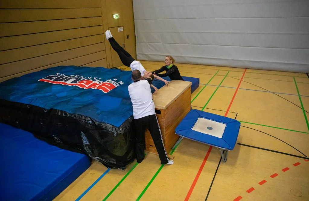 Gymnast flipping onto a blue air pit labeled BAGJUMP as two coaches assist on a gymnasium floor.