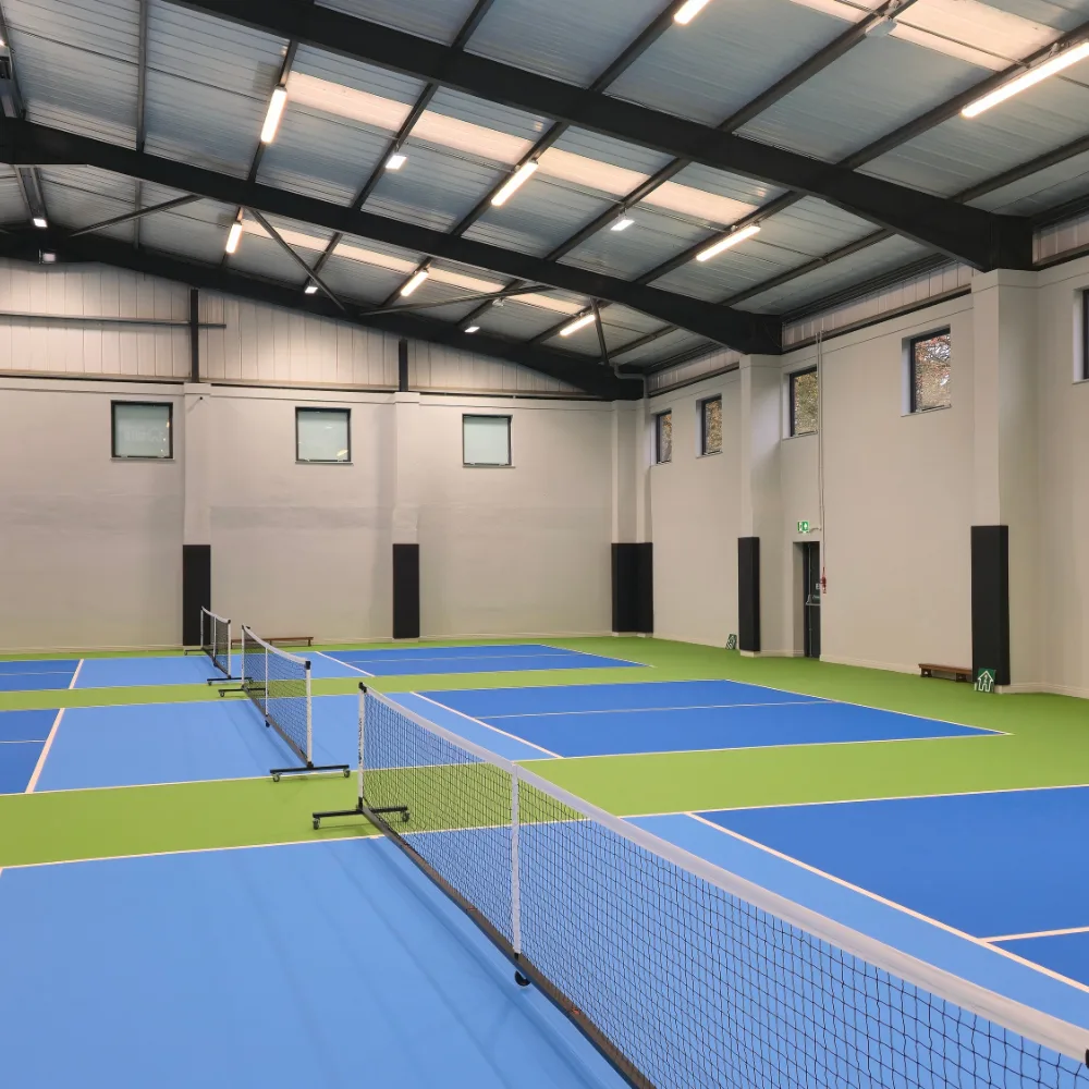 Indoor pickleball courts with blue playing surfaces, green surrounds, portable nets, and black wall padding of high-density foam with PVC covers.