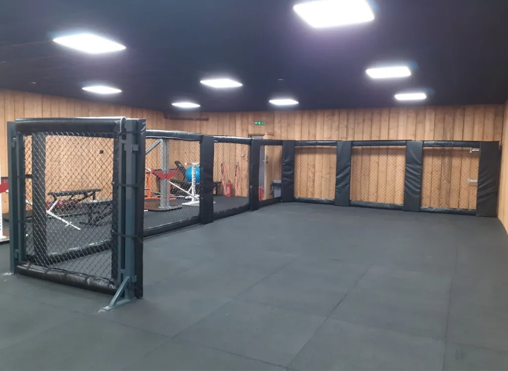 Steel powder-coated cage walling with chain-link sides and black padded corners enclosing a rubber-floored space against wood-paneled walls.