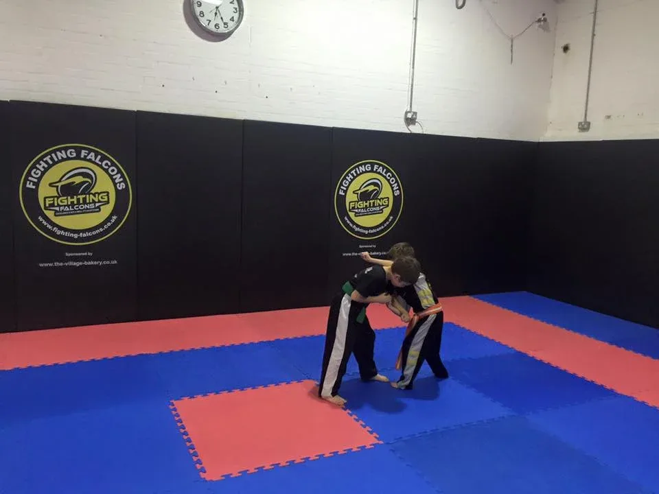 Two children practicing martial arts on reversible red and blue EVA foam jigsaw mats with black padded walls bearing Fighting Falcons logos.