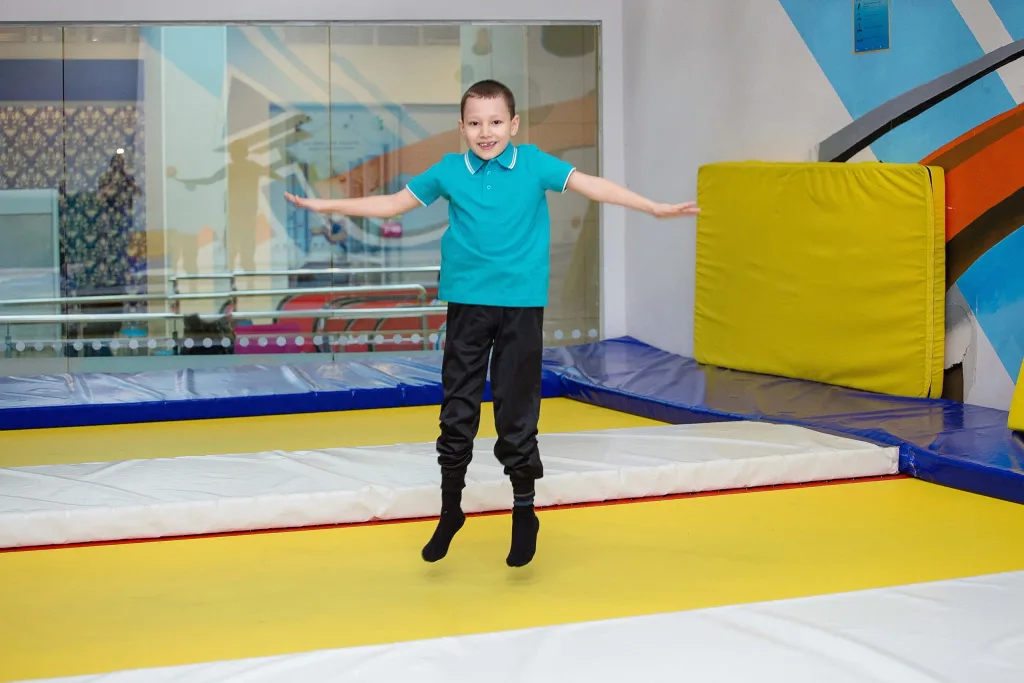 Boy in a teal polo and black pants jumping on a yellow rebound therapy mat with blue edge padding and a green wall pad.