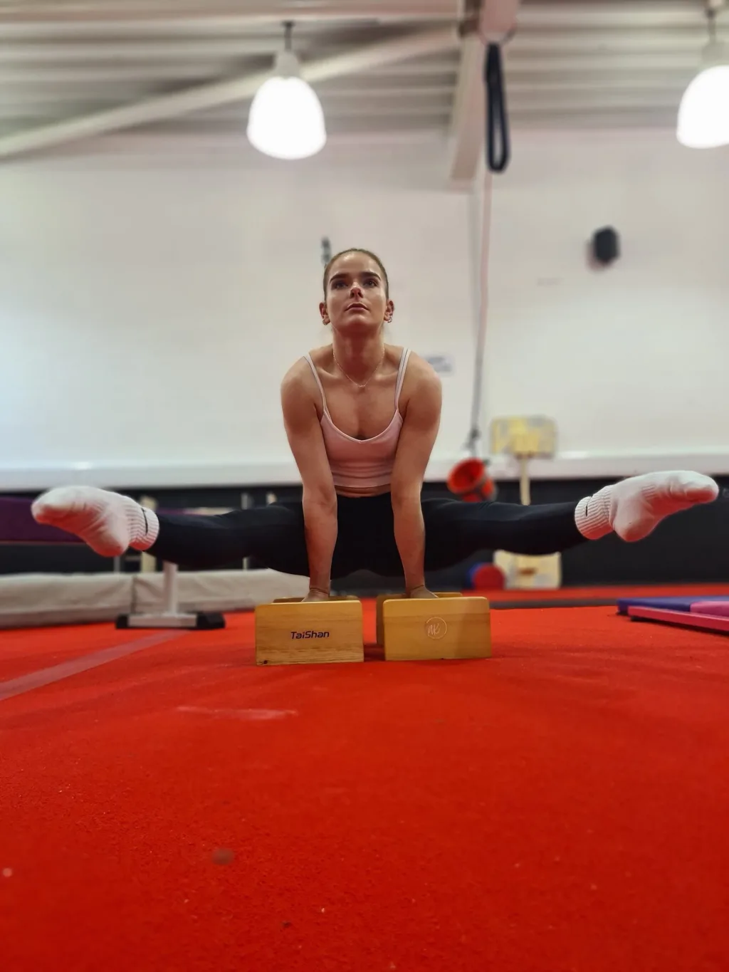 Female gymnast performing a straddle handstand on wooden parallettes labeled TaiShan, on a red gym floor.