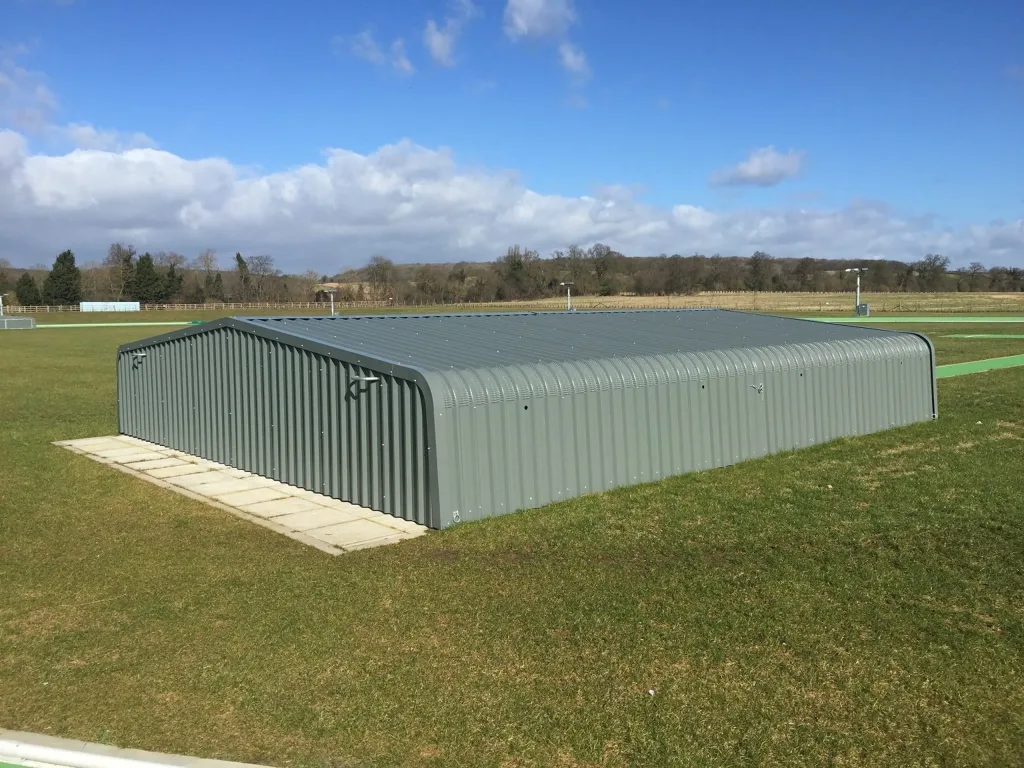 Corrugated galvanised welded steel high jump wheel-away cover with epoxy-coated cladding and jacking handles, lowered flush on a grass field beside a paved strip.