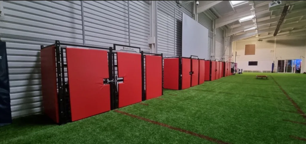 Red PVC sports pitch safety pads lined against a corrugated white wall on green artificial turf inside an indoor sports hall.