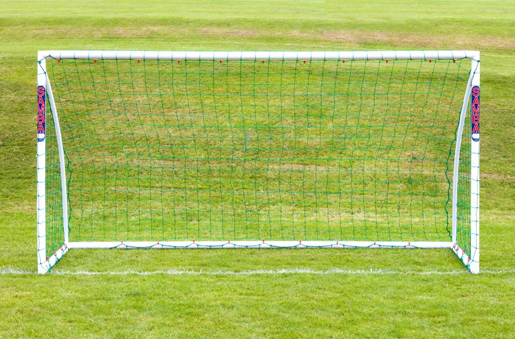White soccer goal with green net and Samba logos on the vertical posts, standing on a grass field.