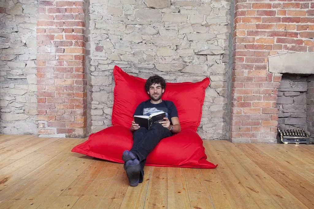 Man sitting and reading on a red cotton floor cushion against a stone and brick wall on a wooden floor.
