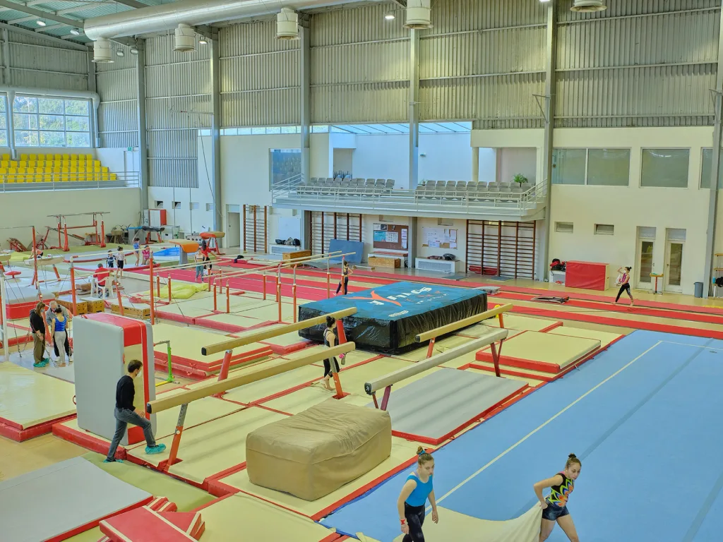 Indoor gymnastics hall with balance beams and red-bordered mats in the foreground and two gymnasts carrying a beige mat on the blue floor.