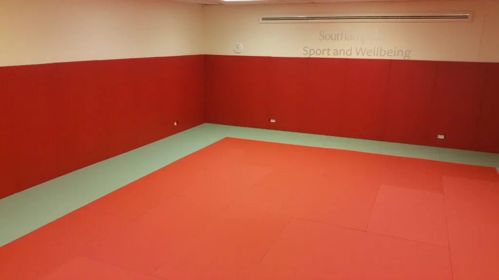 Red tatami-embossed PVC judo mats with a green border in a corner room with red wall pads and wall text 'Southampton Sport and Wellbeing'.
