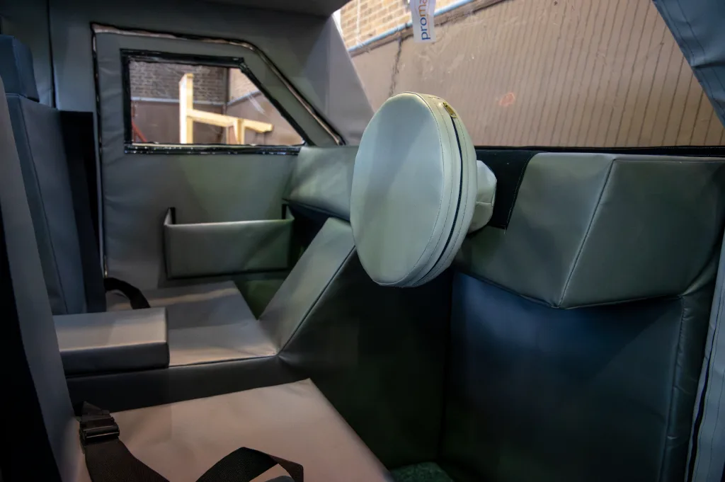 Padded training car interior with grey PVC-covered foam seats, a round cushioned headrest, and a small tag reading "proma".