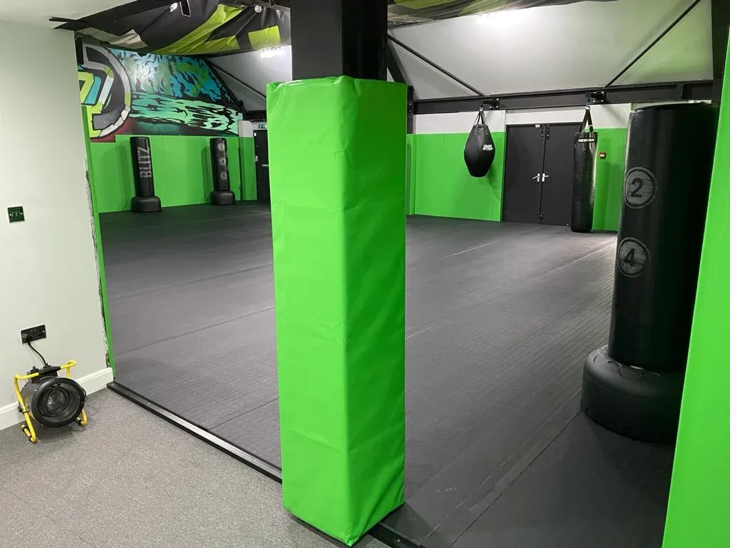 Green padded column protector beside dark roll-out mats in a gym with hanging and freestanding punching bags labeled 2 and 4 and green wall padding.