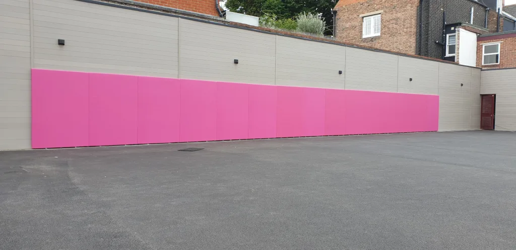 Row of pink PVC-covered wall pads attached to a pale beige exterior wall above an asphalt playground.