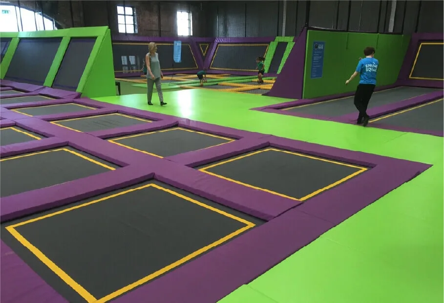 Indoor trampoline park with purple-bordered black trampolines on a green floor and a few people walking.