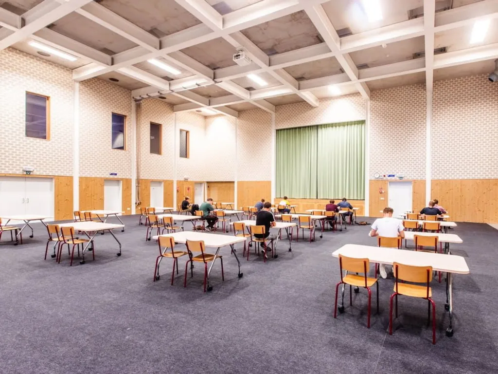 Sports hall carpet tiles cover the floor, with spaced exam tables and chairs and students seated under a high ceiling and green stage curtain.