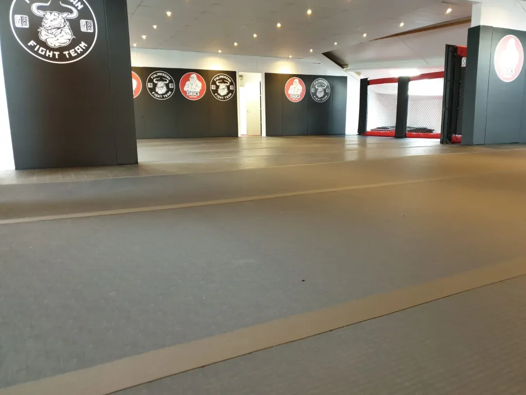 Grey roll-out mats cover a training gym floor with wall logos reading 'FIGHT TEAM' and 'SBG', and a red-trimmed cage at the rear.