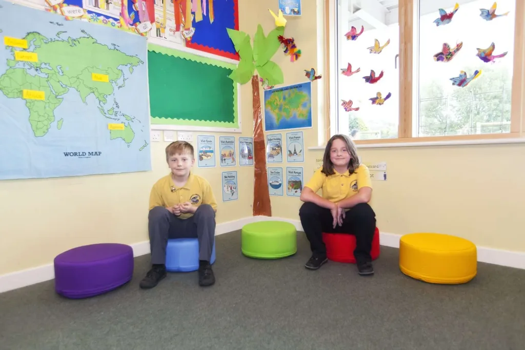 Two children in yellow shirts sit on five round cotton foam seating pods in a classroom with a wall map labeled WORLD MAP.