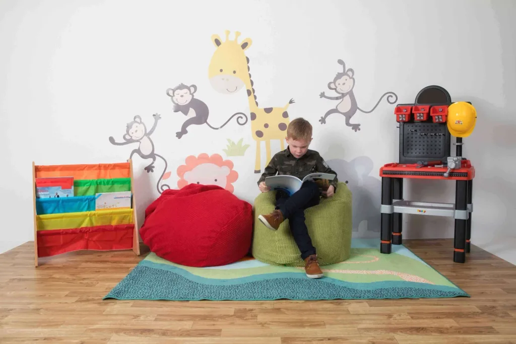 Boy on a green bean bag reading a book, red bean bag beside him, colorful book rack left and toy workbench right, animal wall decals behind.