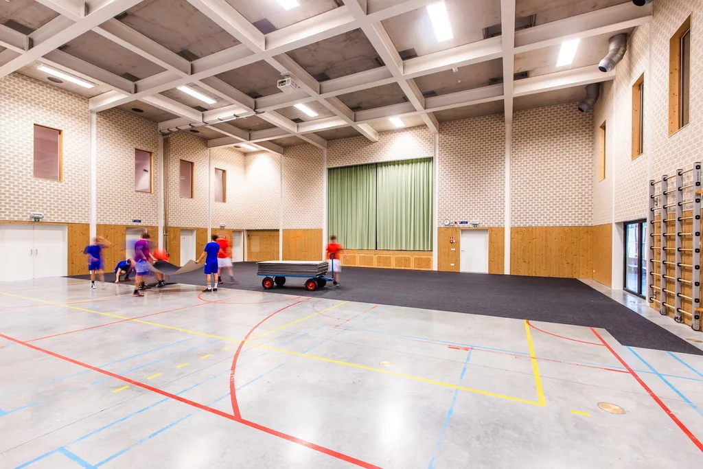 People roll out a dark mat on a gym's concrete court with red, yellow, and blue lines, a green stage curtain, and a cart of mats.